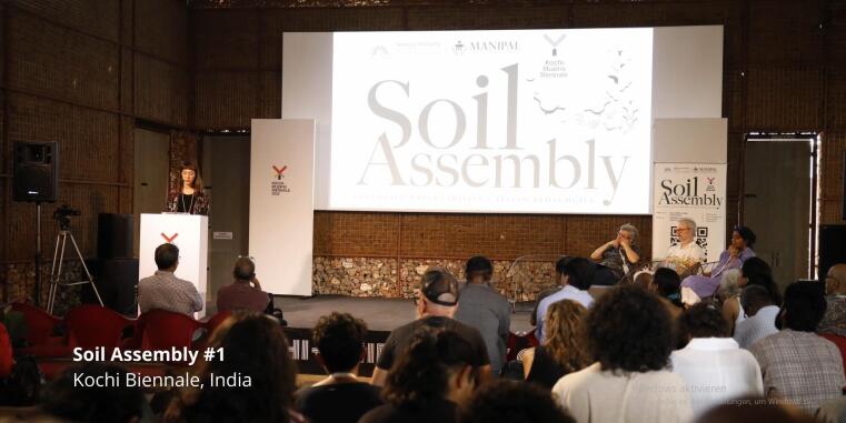 Foto des Veranstaltungsraums, einer vortragenden Frau auf der Kochi Biennale