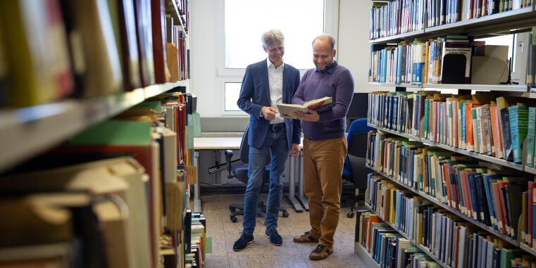 Prof. Dr. Michael Klasen (links) und Dr. Tomáš Ježo vom Institut für Theoretische Physik stehen zwischen zwei Bücherregalen und schauen gemeinsam in ein aufgeschlagenes Buch, das Tomáš Ježo in den Händen hält.