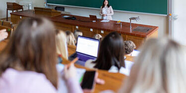 Studierende sitzen mit Laptops in einem Hörsaal und folgen einer Dozentin, die vorne am Pult steht.