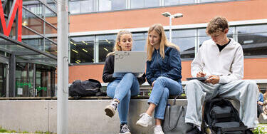 Deutsch: Drei Studierende sitzen draußen auf einer Bank vor einem Universitätsgebäude und arbeiten gemeinsam an einem Laptop.