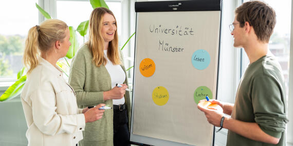 Drei Personen stehen an einem Flipchart mit der Aufschrift „Universität Münster" und farbigen Kärtchen mit den Begriffen „Wissen", „Leben", „Studium" und „Lehre" und diskutieren miteinander.