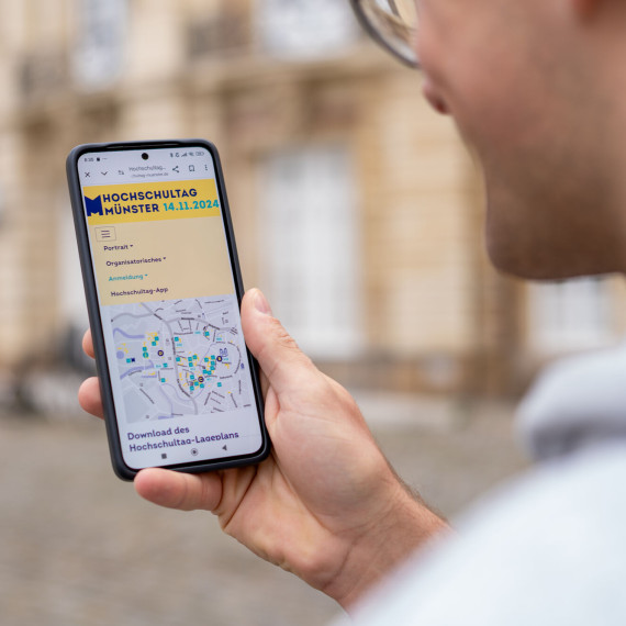 Ein junger Mann hält ein Smartphone in der Hand. Auf dem Smartphone ist ein Lageplan des Hochschultags zu sehen.<address>© Uni MS - Johannes Wulf</address>
