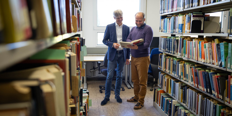 Prof. Dr. Michael Klasen (links) und Dr. Tomáš Ježo vom Institut für Theoretische Physik stehen zwischen zwei Bücherregalen und schauen gemeinsam in ein aufgeschlagenes Buch, das Tomáš Ježo in den Händen hält.<address>© Uni MS - Linus Peikenkamp</address>