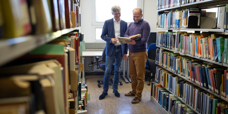 Prof Michael Klasen (left) and Dr Tomáš Ježo from the Institute of Theoretical Physics stand between two bookshelves and look together at an open book that Tomáš Ježo is holding.<address>© Uni MS - Linus Peikenkamp</address>