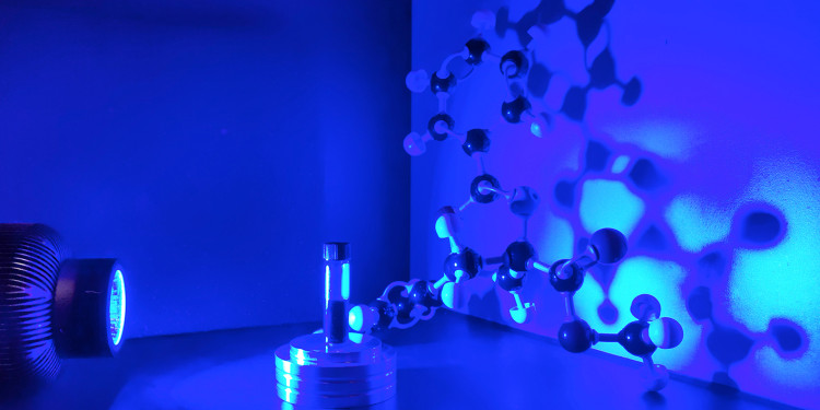 The symbolic photo shows the experimental set-up consisting of a lamp and a glass tube. The figure of a molecule can be seen in the background.<address>© AG Glorius - Dr Chetan Chintawar (symbolic photo)</address>