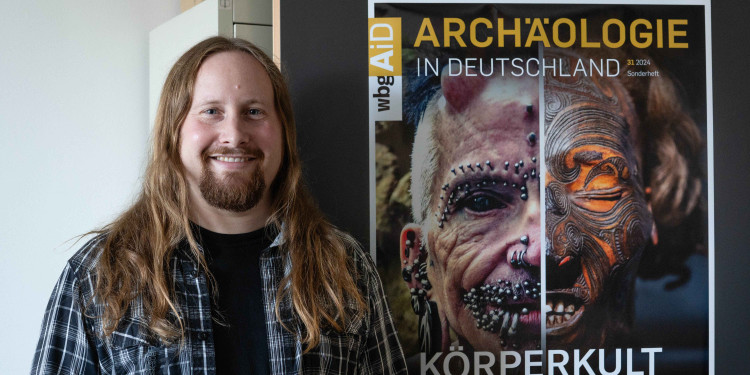 Lukas Kerk vor dem Plakat des Sonderheftes „Körperkult“ der Zeitschrift „Archäologie in Deutschland“.<address>© Uni MS - Linus Peikenkamp</address>