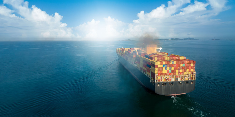 A large container ship is travelling across the sea, emitting a dark cloud of pollutants.<address>© AdobeStock - Yellow Boat</address>