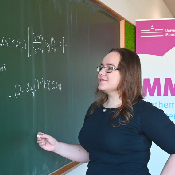 Eine Frau steht vor einer grünen Tafel und schreibt eine mathematische Formel auf.<address>© Uni MS - Victoria Liesche</address>