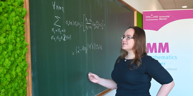 A woman stands in front of a green board and writes down a mathematical equation.<address>© Uni MS - Victoria Liesche</address>