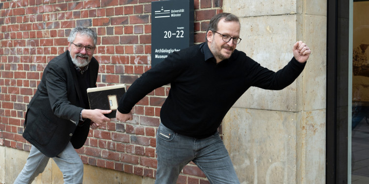 Zu sehen sind Dr. Helge Nieswandt (l.) und sein Nachfolger Dr. Torben Schreiber vor dem Türschild des Archäologischen Museums, die wie bei einem Staffellauf das Inventarbuch in den Händen halten.<address>© Uni MS - Brigitte Heeke</address>