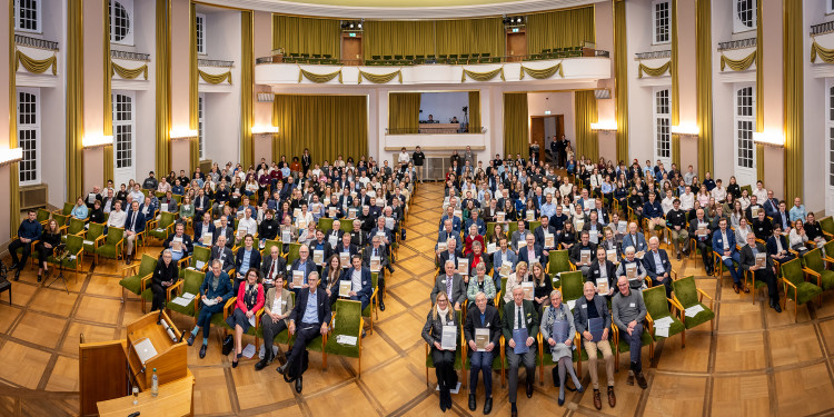 Der Panoramablick von oben in die Aula zeigt Dutzende Stuhlreihen, in denen Hunderte Personen mit Urkunden sitzen und in die Kamera schauen.<address>© Uni MS - Thomas Mohn</address>